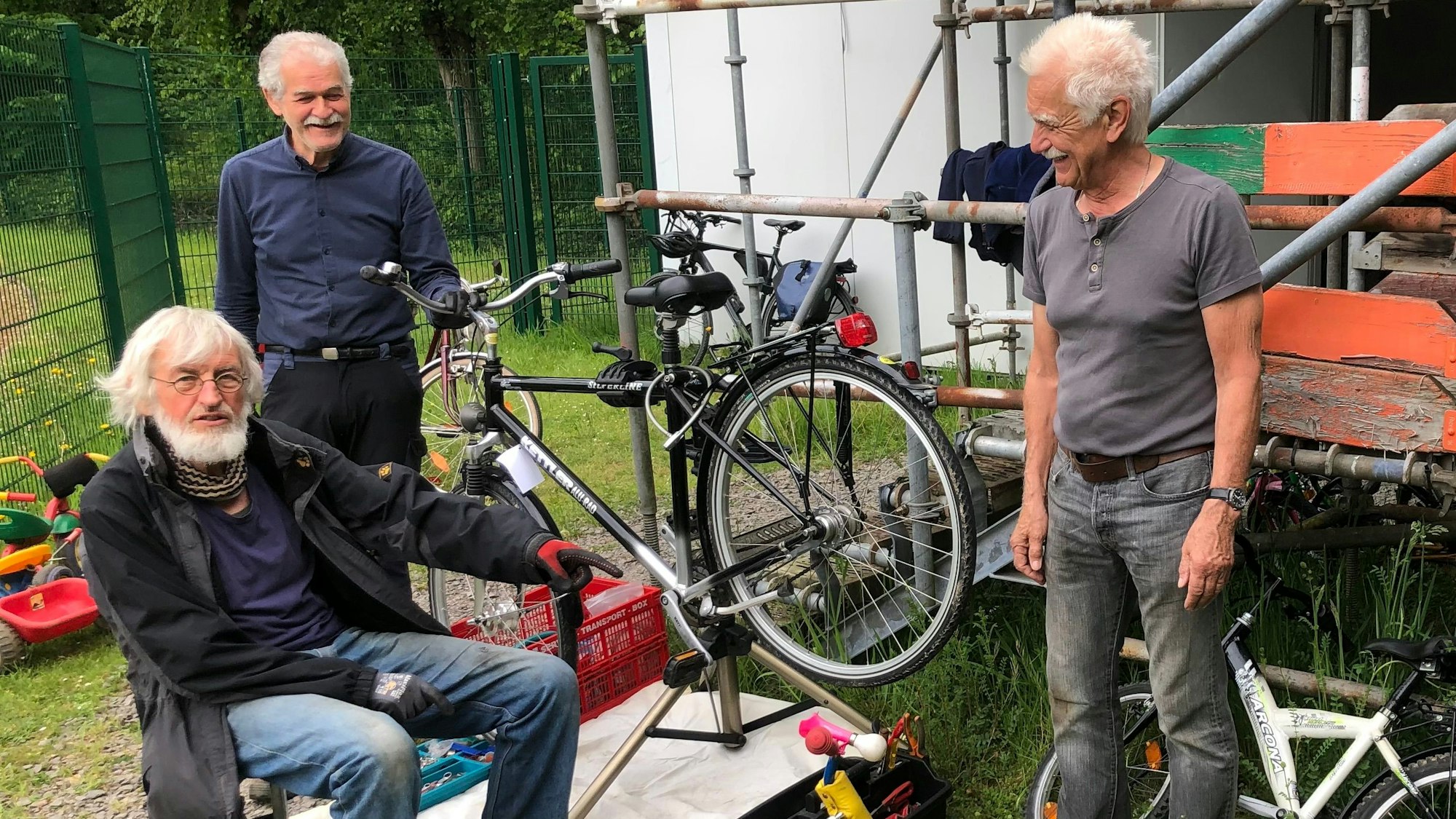 Drei ältere Männer stehen bei einem Fahrrad, das repariert wird, an einem Gerüst.