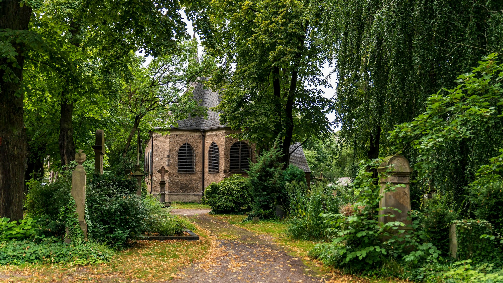 ein Weg führt an Gräbern und grünen Bäumen vorbei zu einem Gebäude