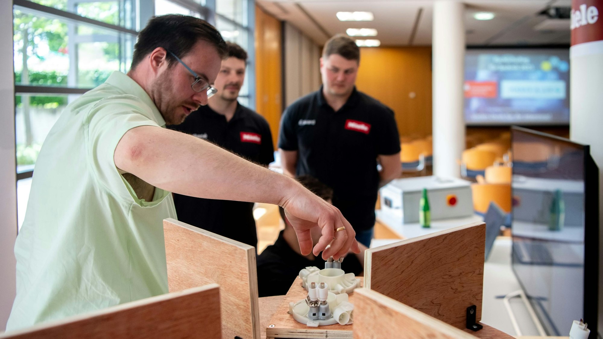 Das Bild zeigt einen Mann, der eine neue Maschine erklärt. Zwei weitere Männer stehen im Hintergrund und schauen zu.