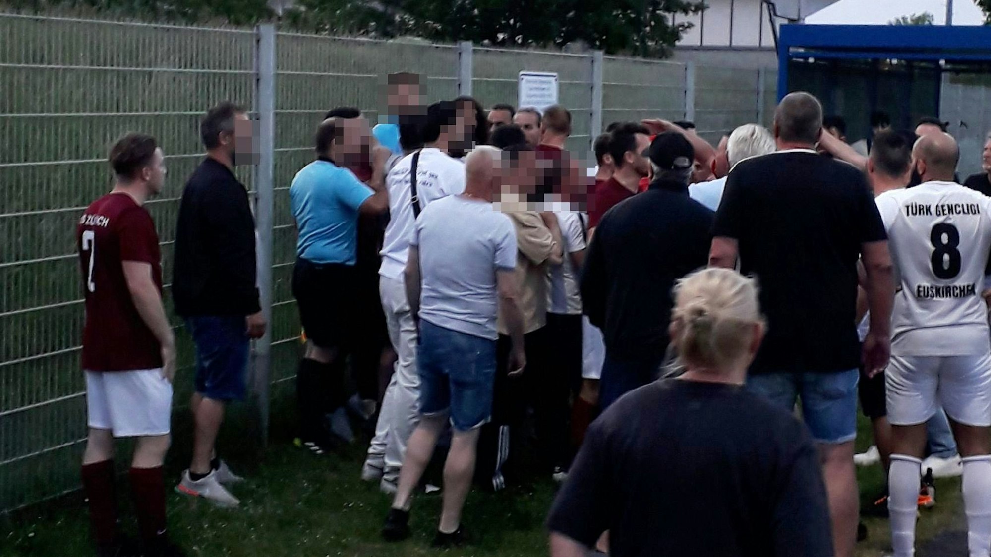 Der Schiedsrichter und ein Assistent stehen am Zaun des Erftstadions, vor ihnen eine aufgebrachte Menge und Menschen, die sie trennen.
