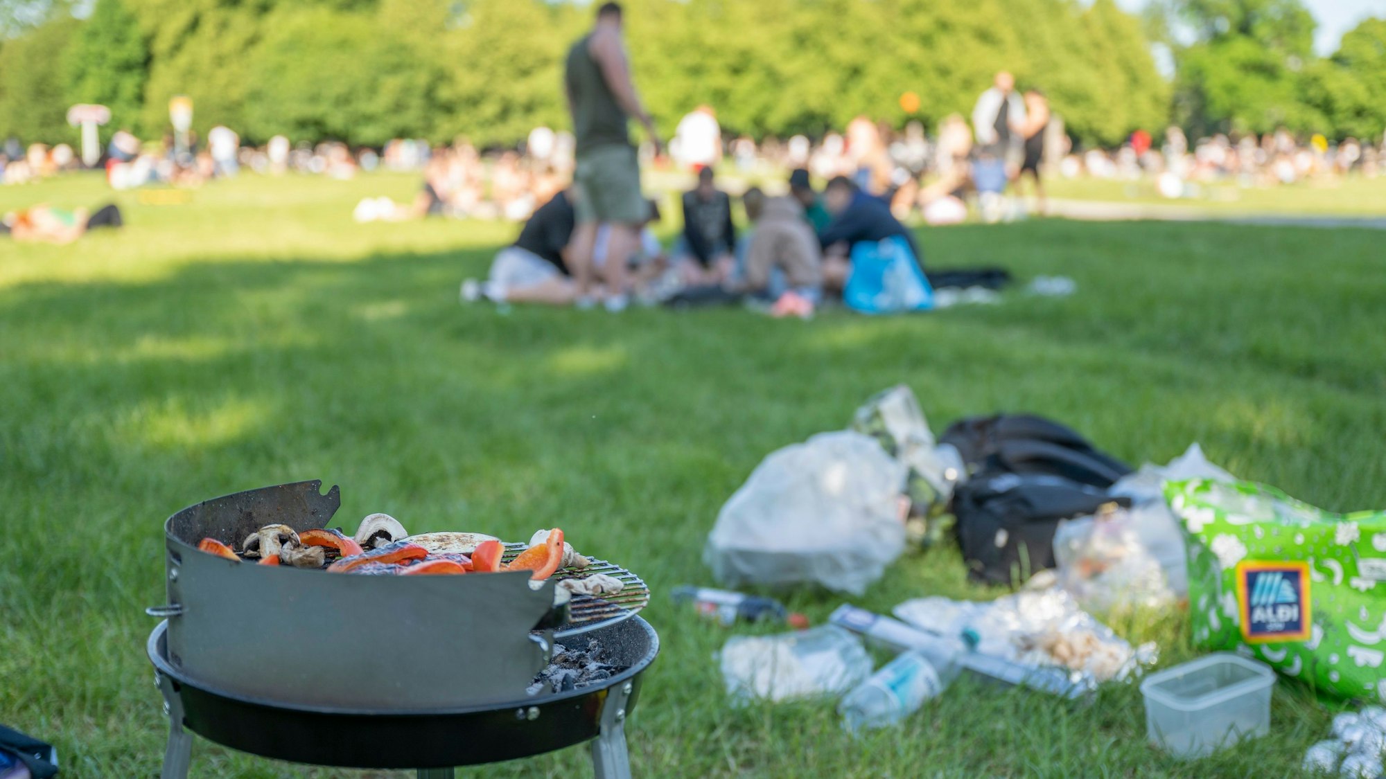 Ein Grill und Tüten und Flaschen im Vordergrund, im Hintergrund sitzen viele Menschen.