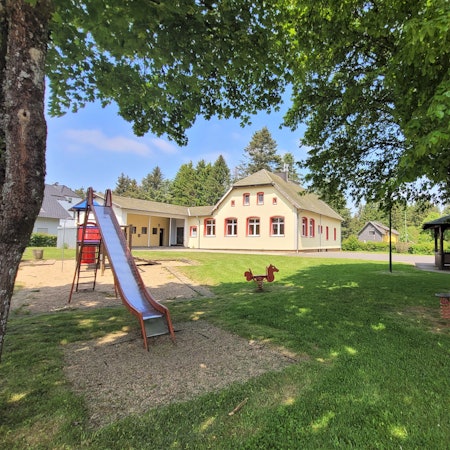 Das Bild zeigt den Dorfplatz in Broich mit einer Rutsche und einem Grillplatz. Im Hintergrund ist das Dorfgemeinschaftshaus zu sehen.