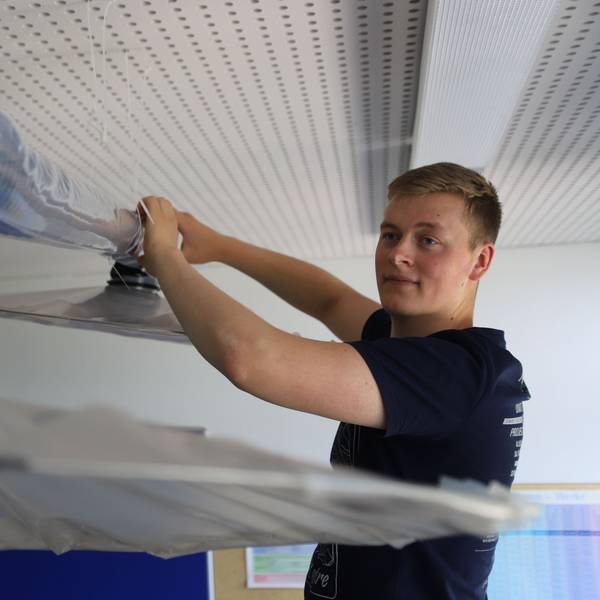 Der Student Bastian Scho greift mit beiden Händen an ein Rohr, das zu einer Lüftungsanlage gehört.