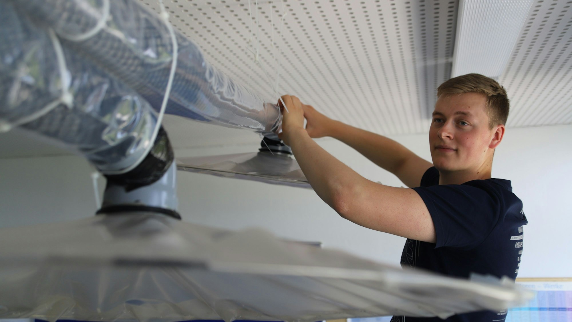 Der Student Bastian Scho greift mit beiden Händen an ein Rohr, das zu einer Lüftungsanlage gehört.
