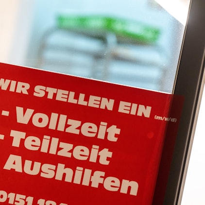 Ein rotes Schild mit der Aufschrift ·„Wir stellen ein - Vollzeit, Teilzeit, Aushilfen“ hängt an einer Eingangstür.