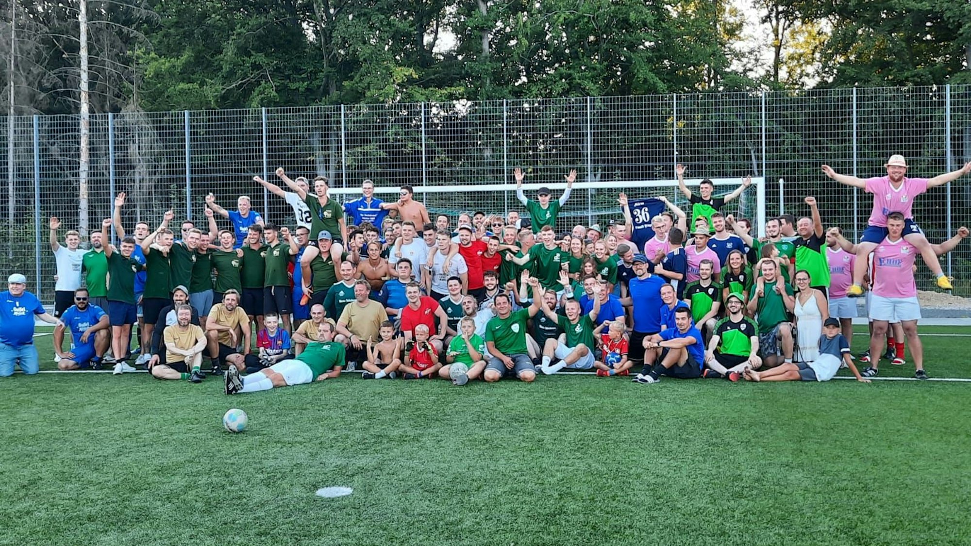 Menschen aller Altersklassen in Fußballtrikots präsentieren sich auf einem Gruppenbild vor einem Fußballtor auf Kunstrasen.