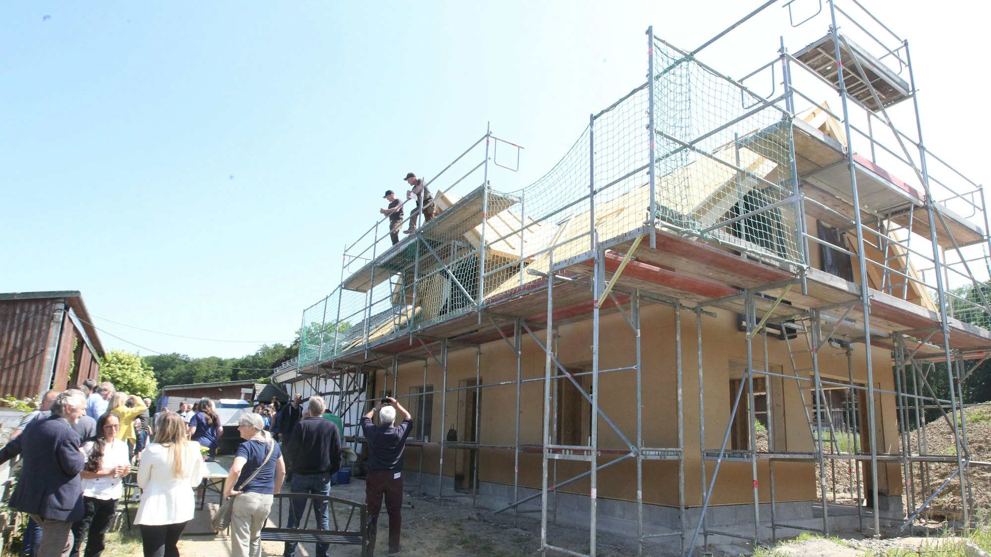 Neben einem Rohbau mit Gerüsten versammeln sich verschiedene Personen.