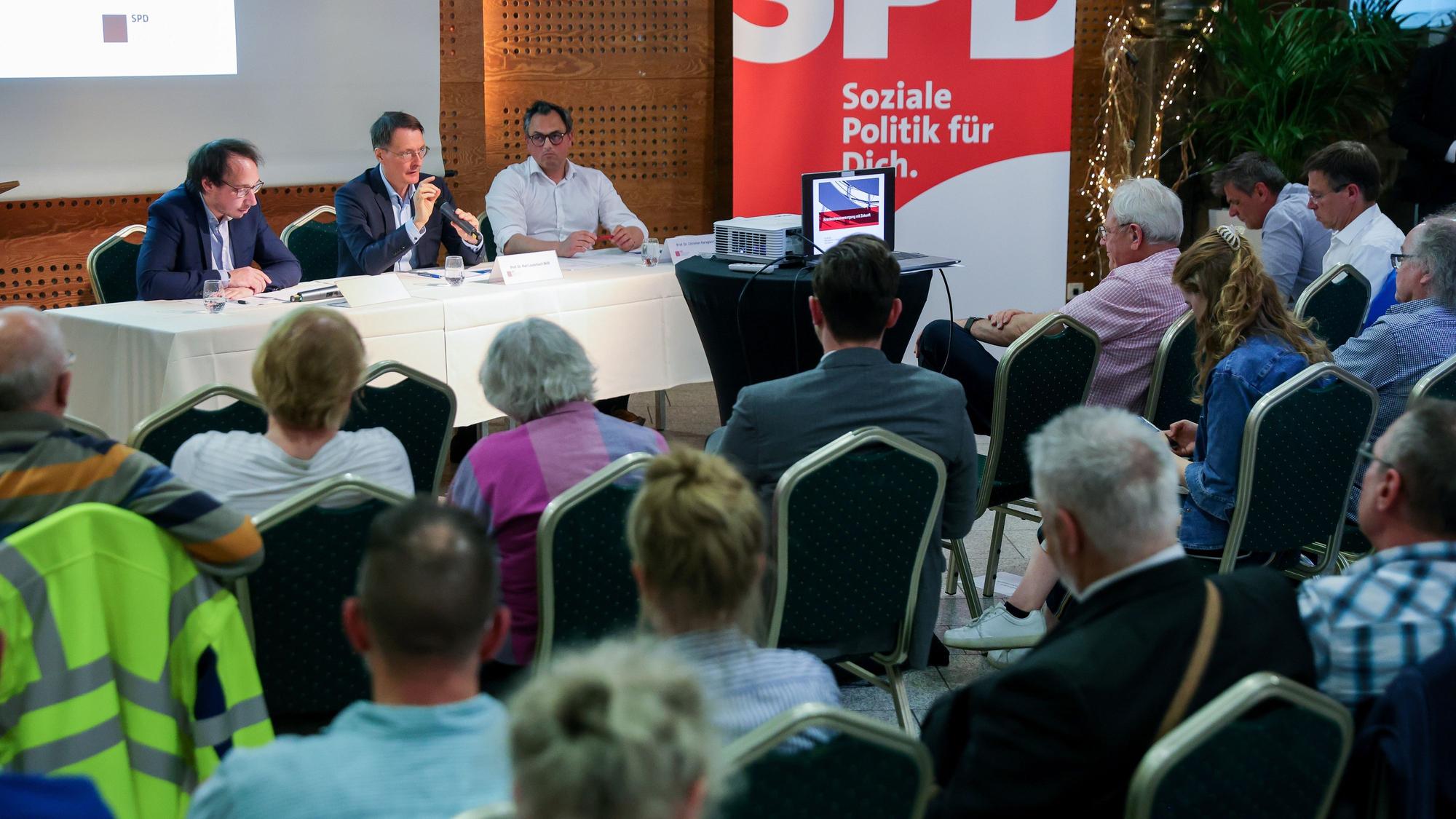 Der Moderator, Prof. Dr. Karl Lauterbach und Prof. Dr. Christian Karagiannidis sitzen auf dem Podium – im Vordergrund sitzen Menschen.