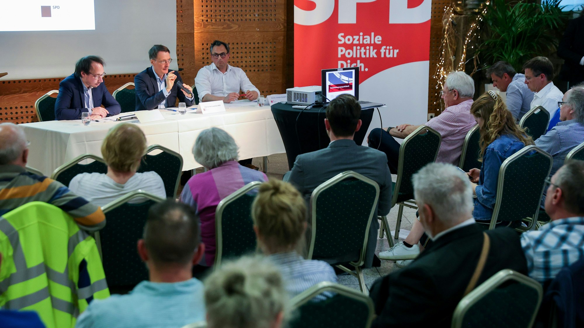 Der Moderator, Prof. Dr. Karl Lauterbach und Prof. Dr. Christian Karagiannidis sitzen auf dem Podium – im Vordergrund sitzen Menschen.
