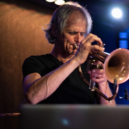 Markus Stockhausen spielt Trompete, er hat die Augen geschlossen. Im Hintergrund eine nicht verschwommene Aufschrift, die blau erleuchtet ist.