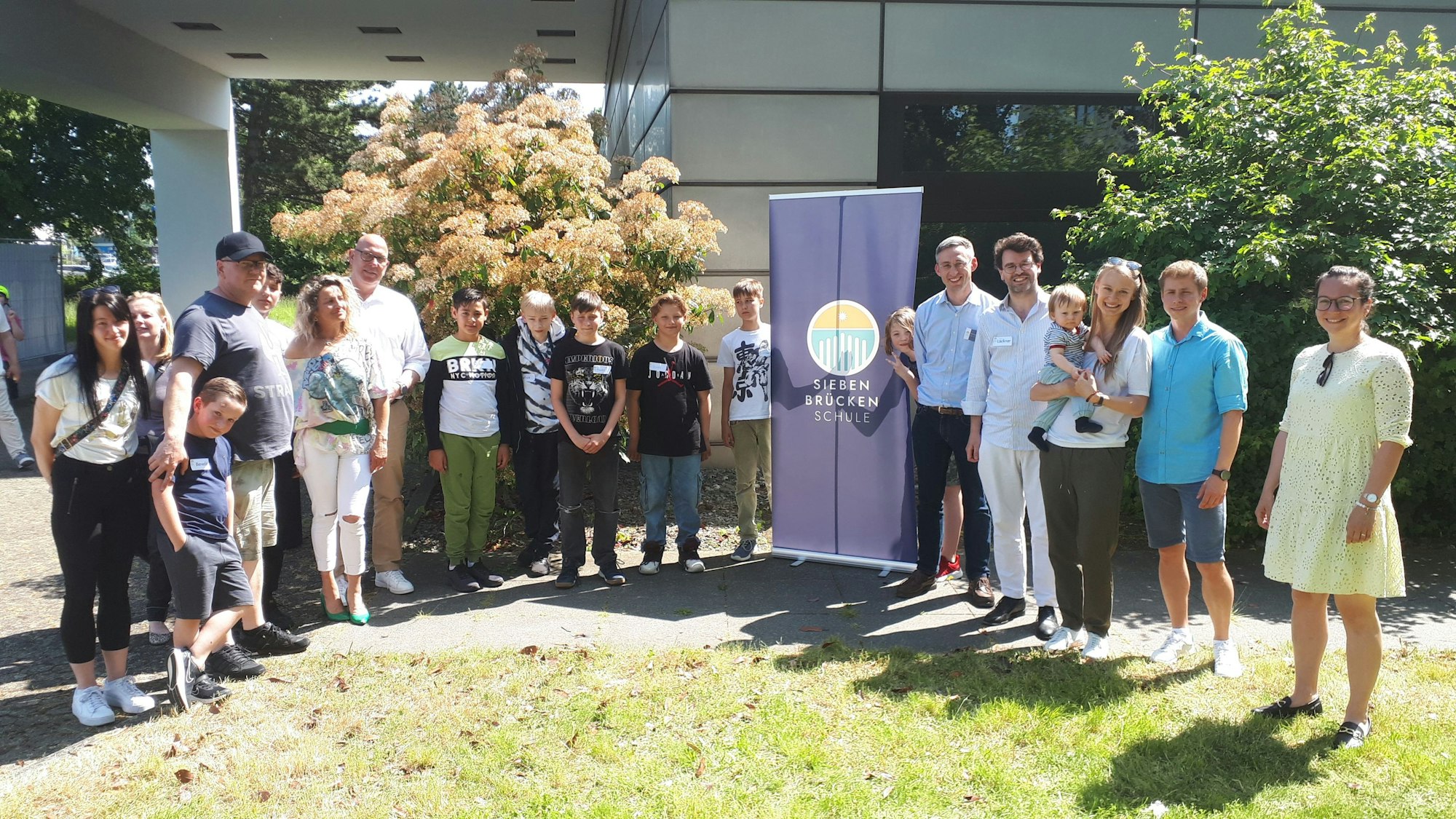 Jugendliche und Erwachsene stehen im Halbkreis auf einem Rasen vor einem Gebäude.