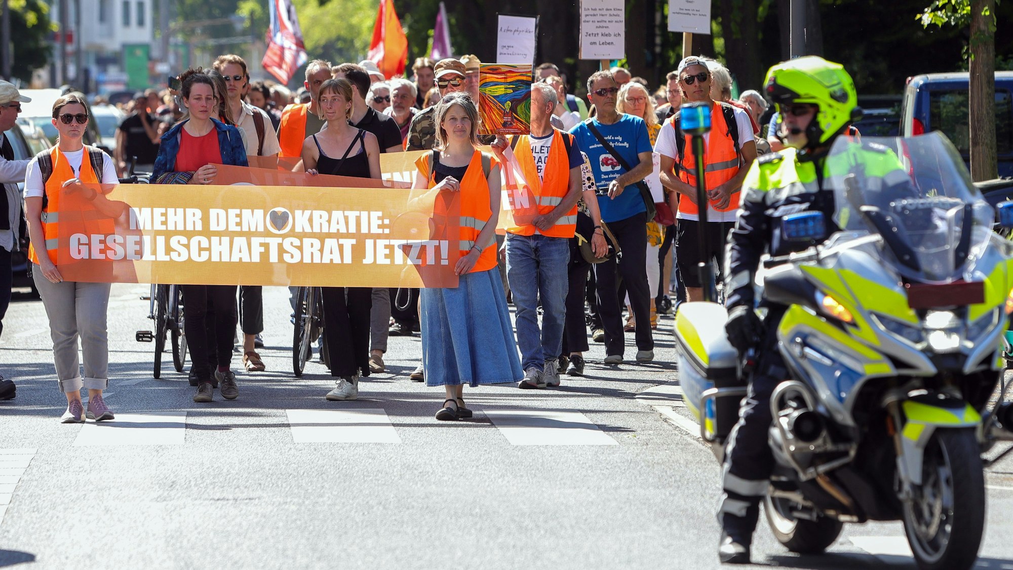 Auf der Straße marschierende Jugendliche halten ein Banner mit der Aufschrift „Mehr Demokratie: Gesellschaftsrat jetzt!“ Ihnen folgen zahlreiche Fußgänger und Radfahrer mit Plakaten und Fahnen. Vor den Marschierenden ist rechts im Bild ein Motorradpolizist zu sehen.