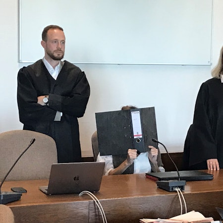 Die Angeklagte sitzt mit einem Ordner vor dem Gesicht auf einem Stuhl, ihre Verteidiger stehen neben ihr.