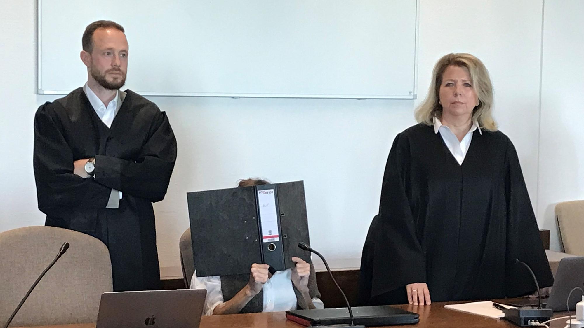 Die Angeklagte sitzt mit einem Ordner vor dem Gesicht auf einem Stuhl, ihre Verteidiger stehen neben ihr.