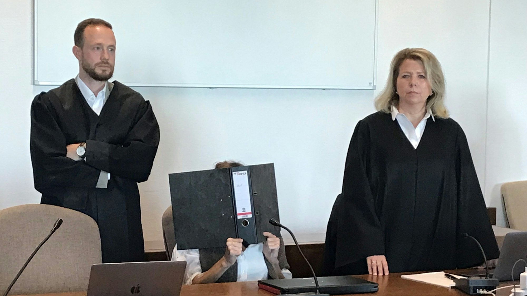 Die Angeklagte sitzt mit einem Ordner vor dem Gesicht auf einem Stuhl, ihre Verteidiger stehen neben ihr.