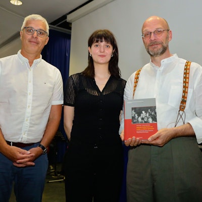 Ein Lehrer hält ein Buch in die Kamera. Daneben stehen ein weiterer Lehrer sowie eine ehemalige Schülerin.