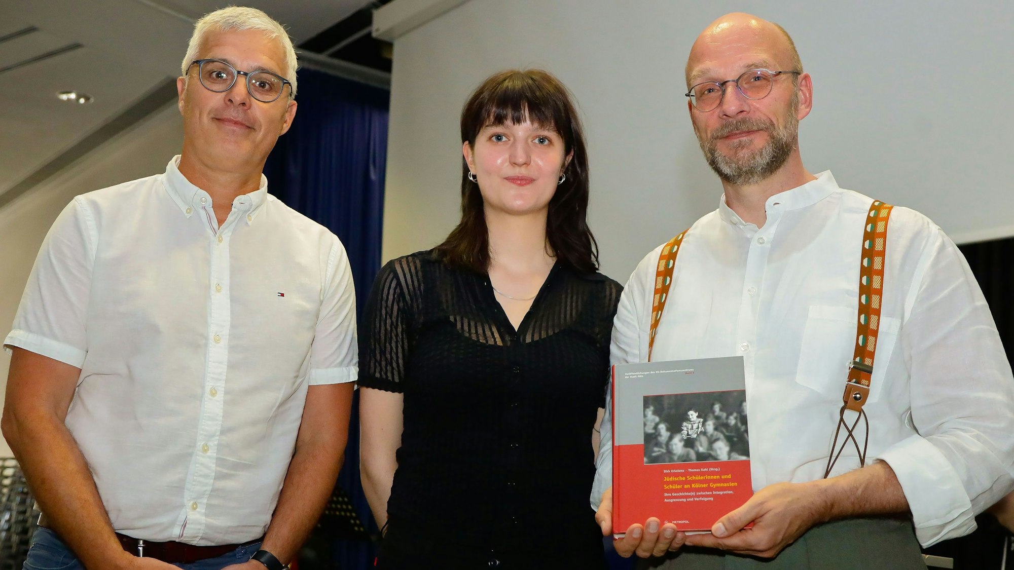 Ein Lehrer hält ein Buch in die Kamera. Daneben stehen ein weiterer Lehrer sowie eine ehemalige Schülerin.