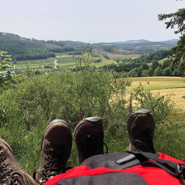 Ein Rucksack und zwei Paar Schuhe stehen vor einer Aussicht des Rothaarsteigs.