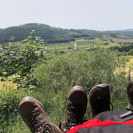 Ein Rucksack und zwei Paar Schuhe stehen vor einer Aussicht des Rothaarsteigs.