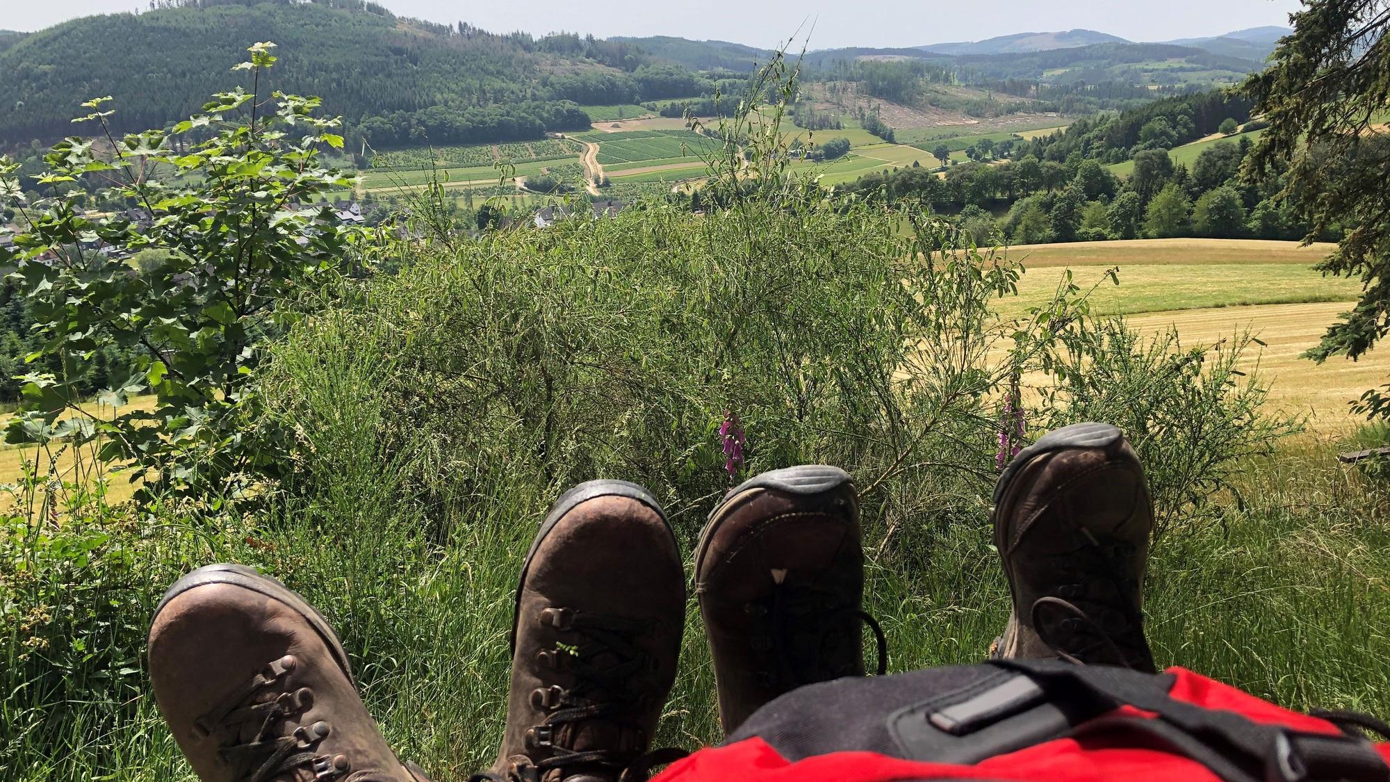 Ein Rucksack und zwei Paar Schuhe stehen vor einer Aussicht des Rothaarsteigs.