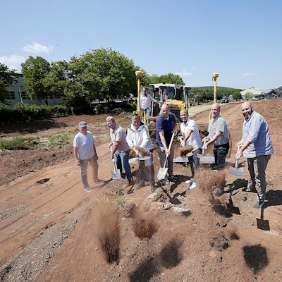 Sieben Männer stehen mit Spaten in der Hand vor einer Baumaschine auf der Erde, wo nun ein Kunstrasenplatz entsteht.