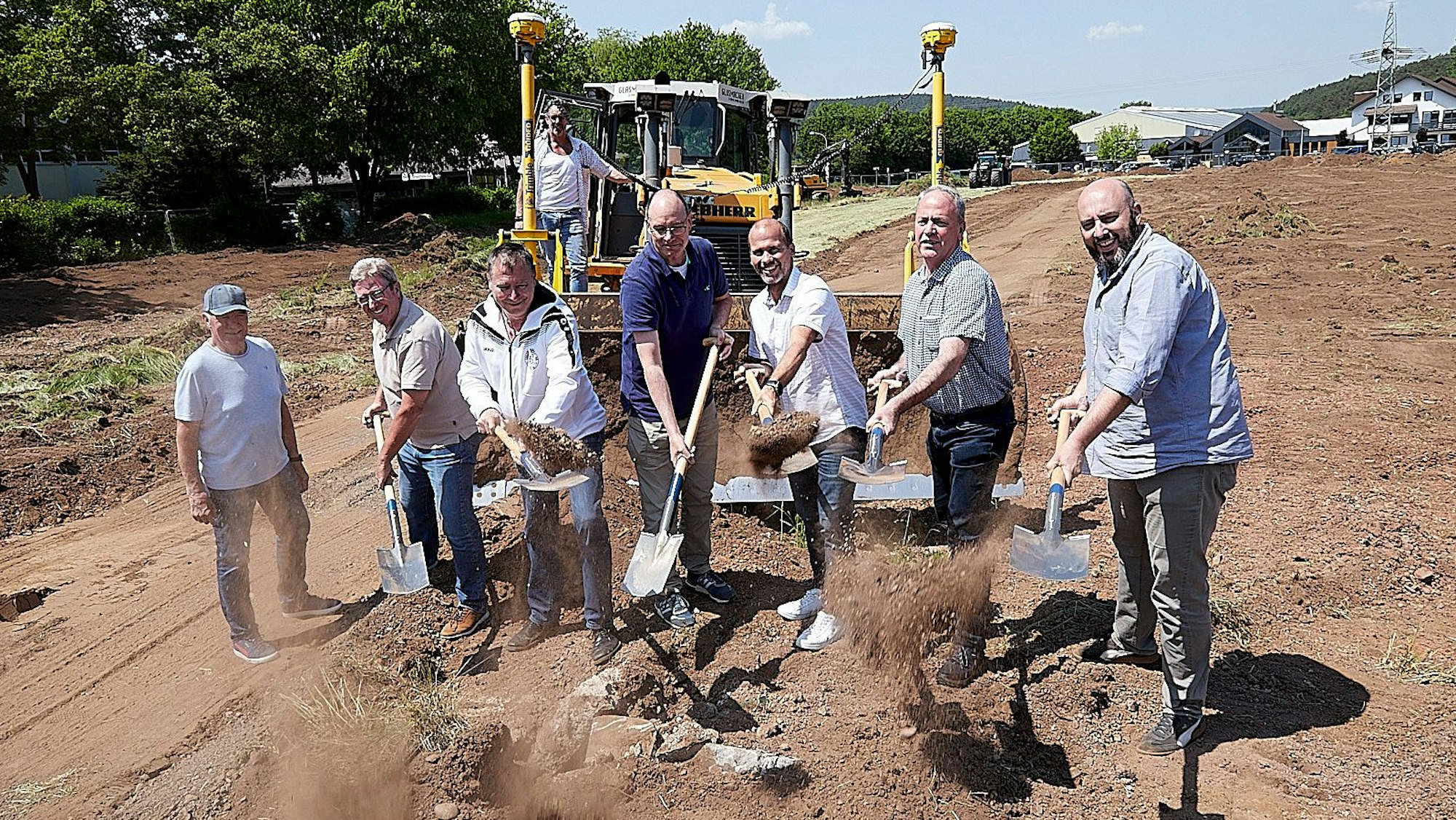 Sieben Männer stehen mit Spaten in der Hand vor einer Baumaschine auf der Erde, wo nun ein Kunstrasenplatz entsteht.
