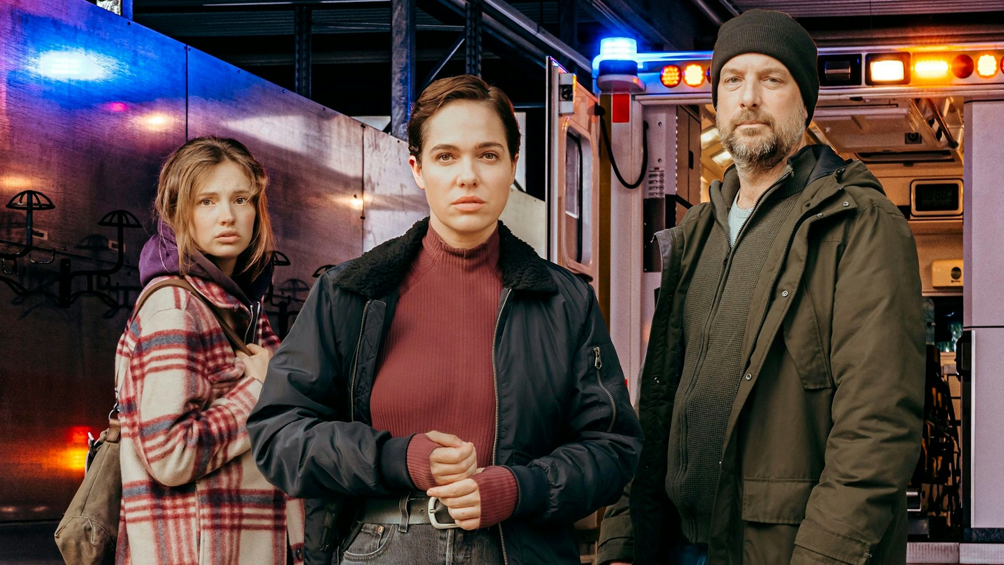 Die drei Schauspieler stehen leicht versetzt nebeneinander und schauen in die Kamera. Hinter ihnen steht ein Krankenwagen.