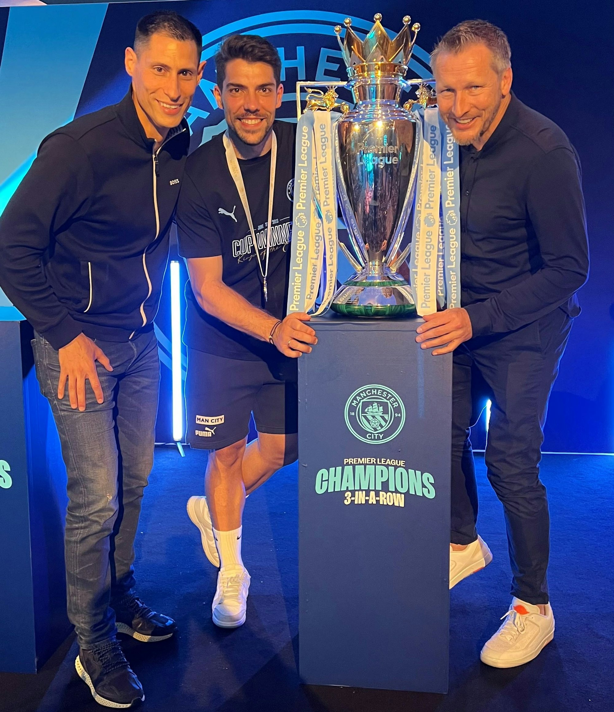 Stefan Ortega (M.), deutscher Torhüter von Manchester City, posiert mit seinen Kölner Beratern Jörg Neblung (r.) und Daniel Valdivieso (l.) mit dem engischen Meisterpokal.