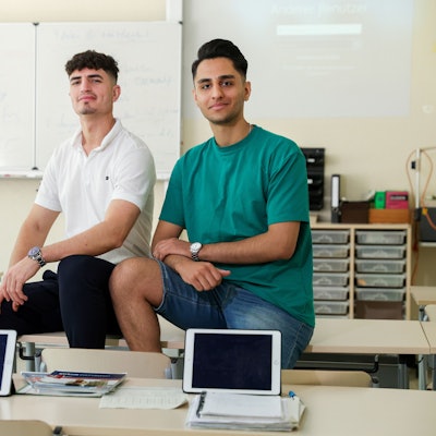 Zwei junge Männer sitzen in einer Schulklasse des Berufskollegs Südstadt auf einem Pult.