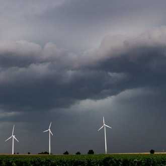 Eine Unwetterfront und dunkle Wolken ziehen über den Himmel. Davor ragen fünf Windräder in den Himmel.