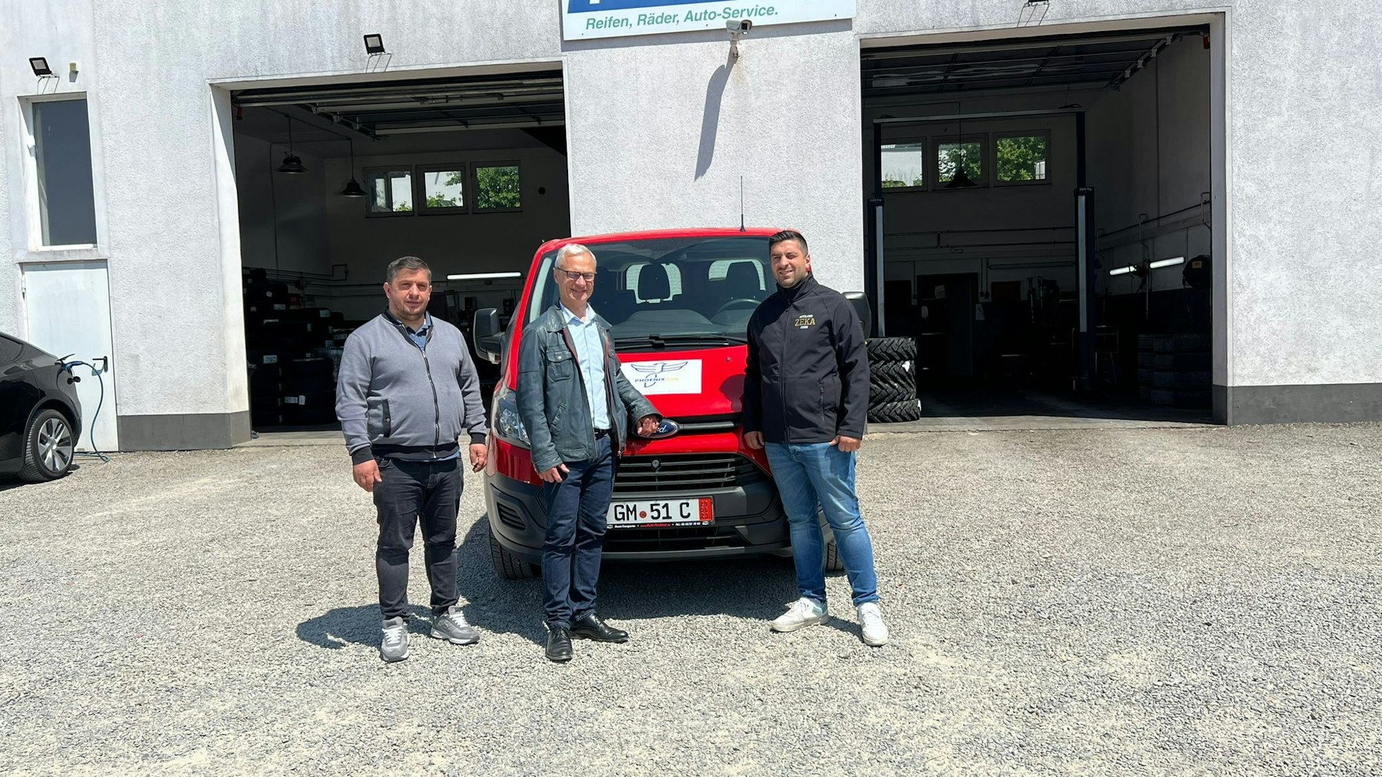 Mittig vor zwei geöffneten Garagentoren steht ein roter Lieferwagen. Vor diesem posieren drei Männer für die Kameraaufnahme.