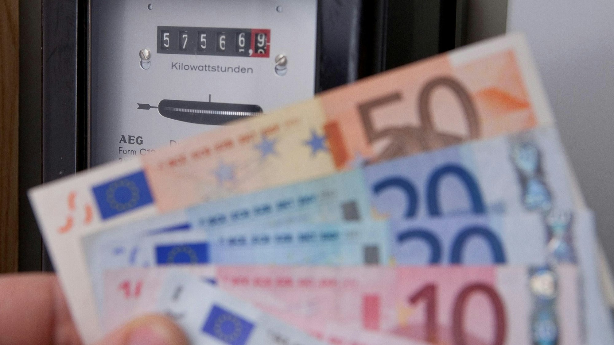 Eine Person hält Geldscheine in der Hand, im Hintergrund ist ein Stromzähler zu sehen.