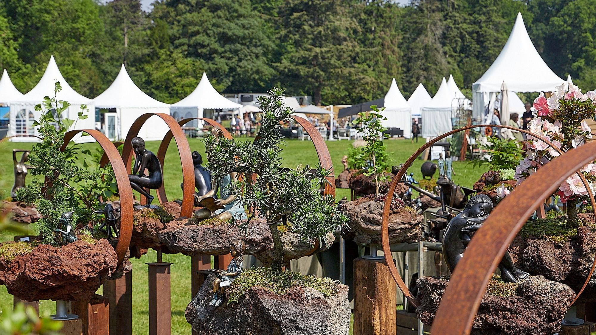 Das Bild zeigt im Vordergrund Kreationen mit Bonsaipflanzen und Metall, im Hintergrund die weißen Zelte der Aussteller der Messe Gartenträume im Garten von Schloss Eicks.