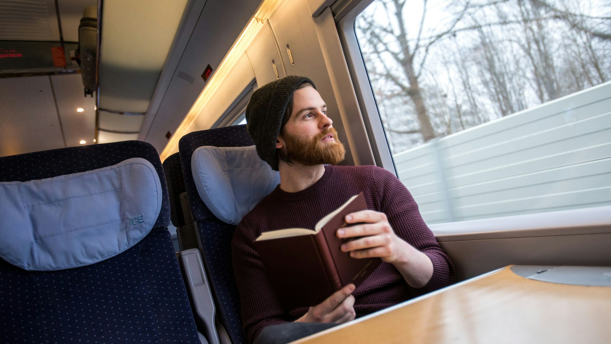 Ein junger Mann sitzt an einem Tisch im ICE. Er hält ein Buch in der Hand und schaut aus dem Fenster.
