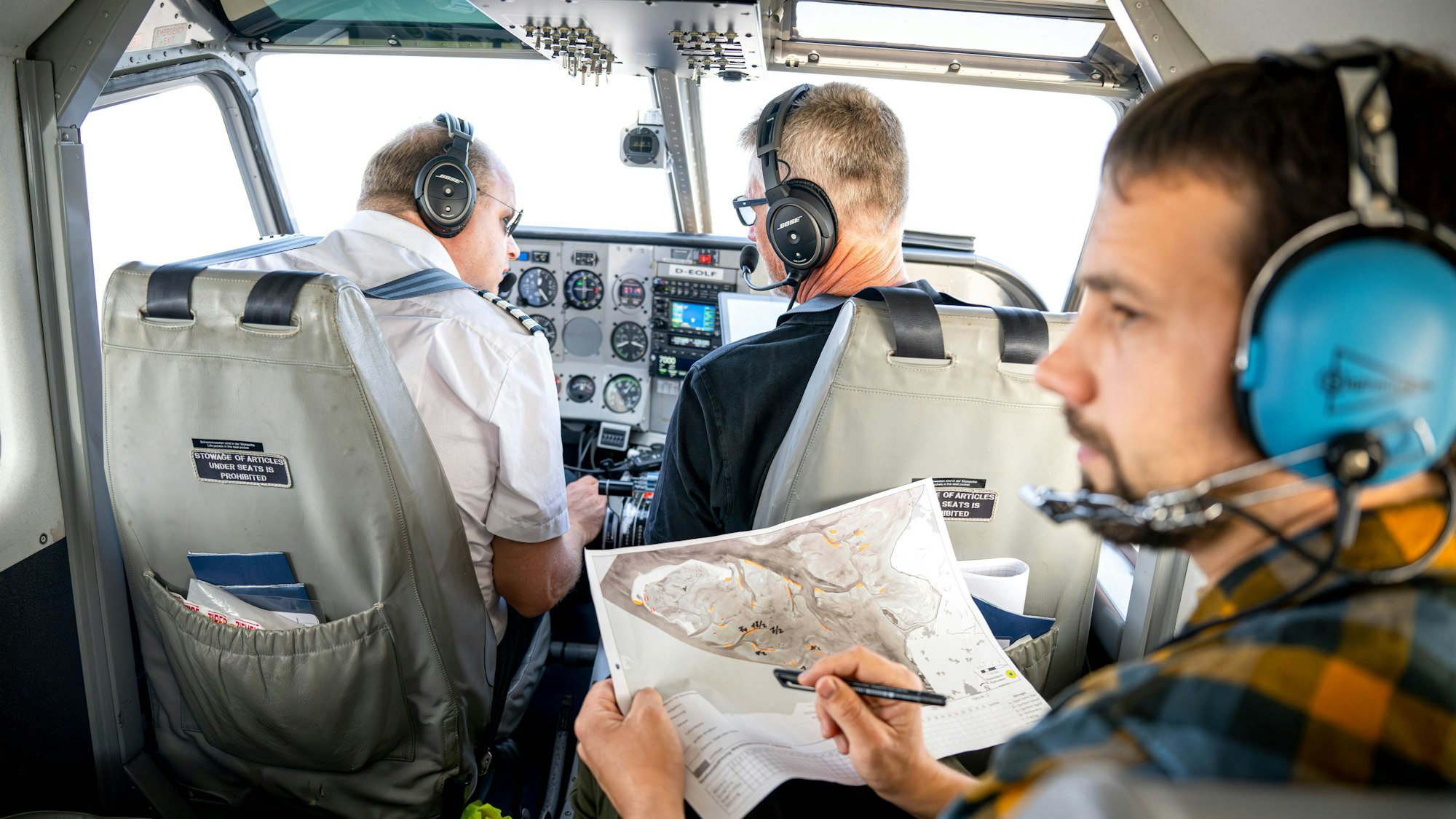 Blick in das Flugzeug. Pilot und Co-Pilot schauen nach vorne. Auf einem Sitz hinter ihnen hält Ole Stejskal eine Karte in der Hand und schaut nach links.