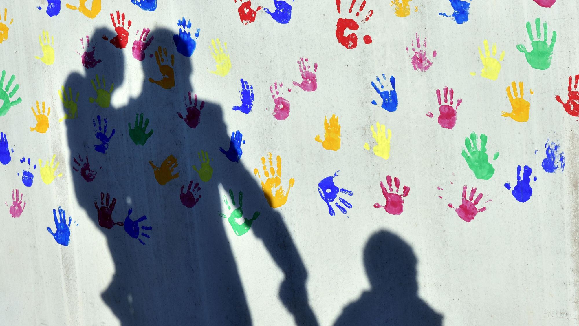 Ein Mann mit einem Kind auf dem Arm und einem an der Hand wirft einen Schatten auf eine mit bunten Handabdrücken bemalte Wand einer Kindertagesstätte. (Illustration zu dpa: «Zu wenige Pflegefamilien in sächsischen Großstädten») +++ dpa-Bildfunk +++