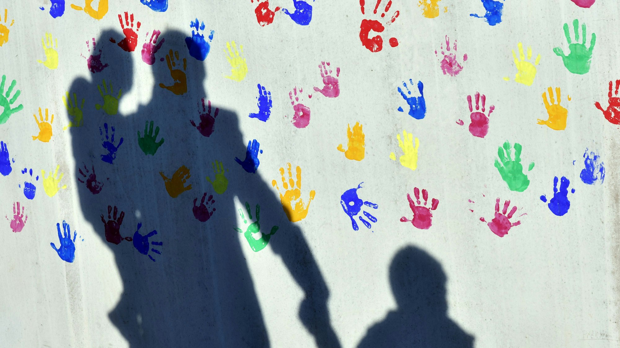 Ein Mann mit einem Kind auf dem Arm und einem an der Hand wirft einen Schatten auf eine mit bunten Handabdrücken bemalte Wand einer Kindertagesstätte. (Illustration zu dpa: «Zu wenige Pflegefamilien in sächsischen Großstädten») +++ dpa-Bildfunk +++