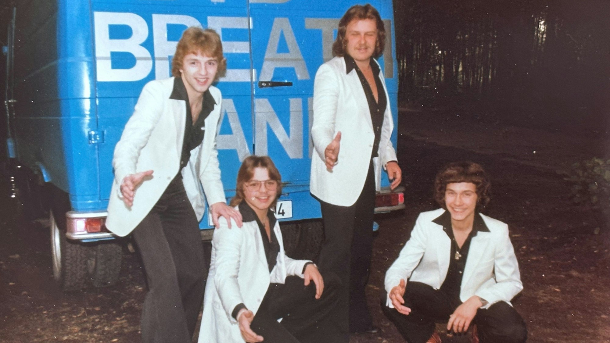 Die vier vor einem blauen Transporter in schwarzen Hemden und Hosen, braunen Lederschuhen und weißem Sakko.