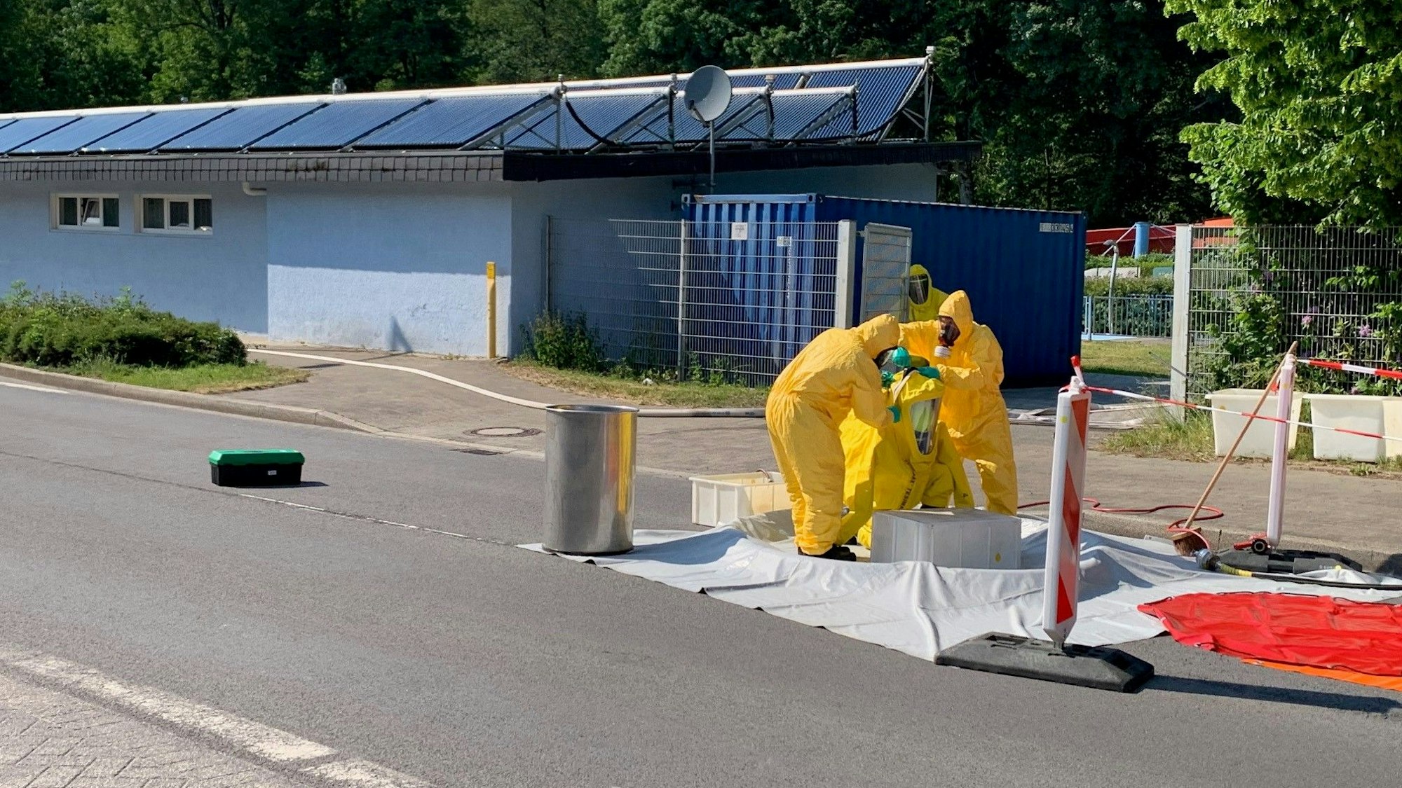 Drei Personen in gelben Ganzkörperanzügen stehen auf einer Straße vor einem Freibad.