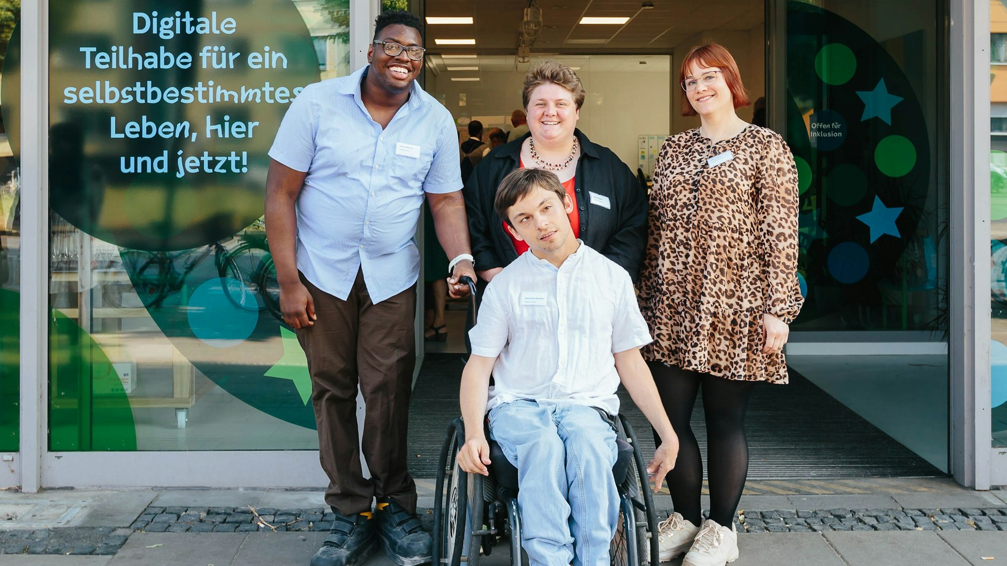 Die vier stehen vor einer geöffneten Eingangstüre, die links den Schriftzug „Digitale Teilhabe für ein selbstbestimmtes Leben, hier und jetzt!“ trägt.