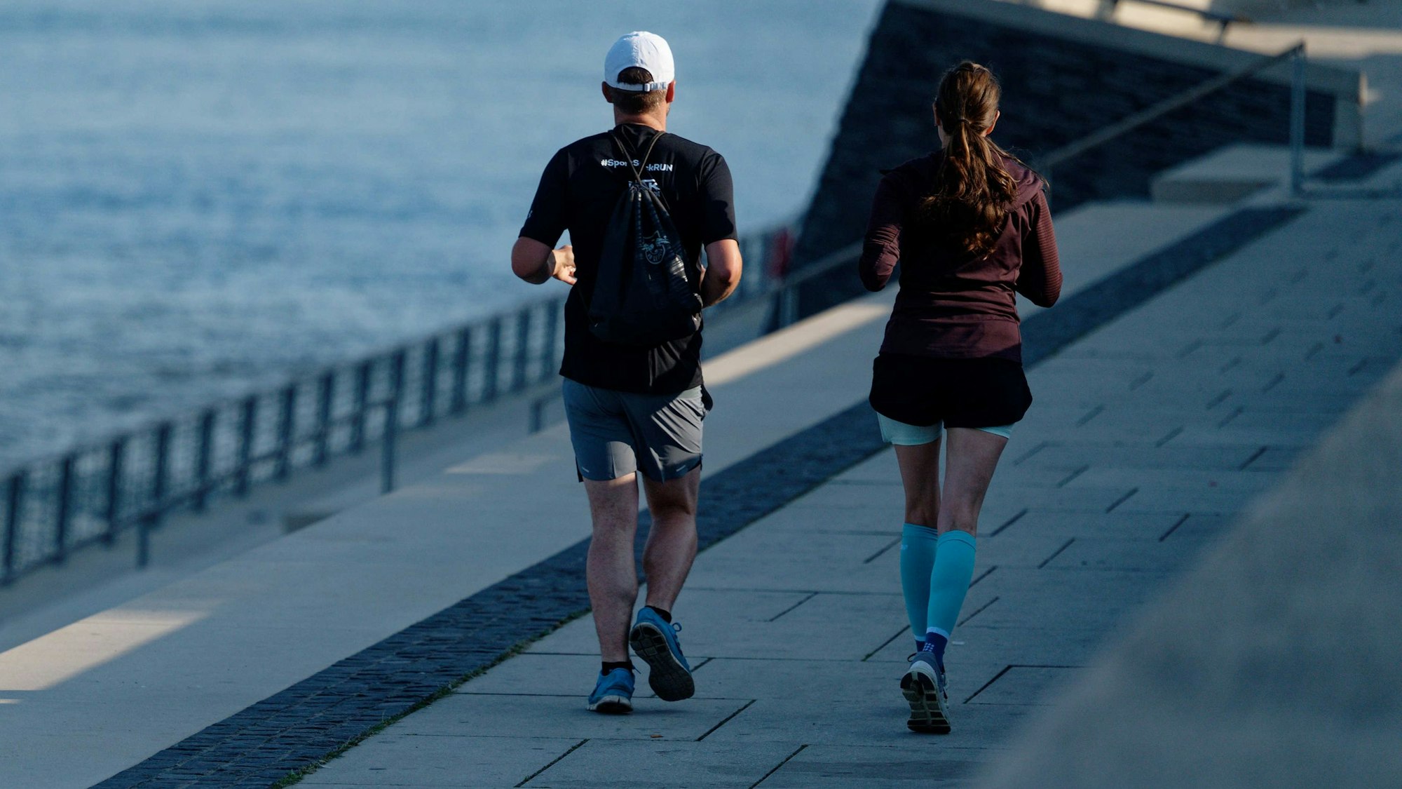 Zwei Jogger laufen in der frischen Morgenluft auf der Rheinpromenade am Rhein entlang.