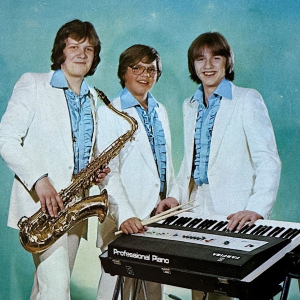 Die drei Bandmitglieder tragen blaue Rüschenhemden und weiße Anzüge.