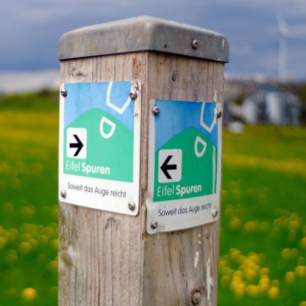 Ein Wegweiser der Eifelspur „Soweit das Auge reicht“ bei Mechernich-Lorbach. Im Hintergrund sind eine Wiese mit gelbem Löwenzahn und ein Windrad zu erahnen.