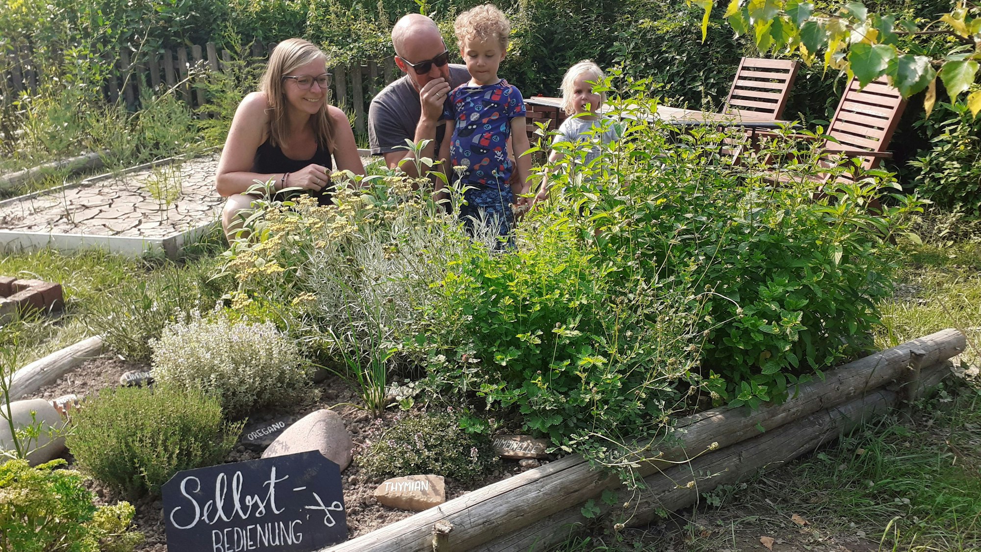 Vor einem Beet mit vielen teils hüfthohen Kräutern steht ein Schild mit der Aufschrift „Selbstbedienung“. Hinter dem Beet hockt eine vierköpfige Familie mit zwei Kindern.
