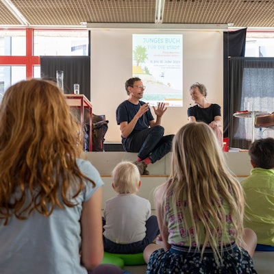 Philip Waechter sitzt mit Moderatorin Britta Weyers auf einer Bühne, davor sitzen Kinder.