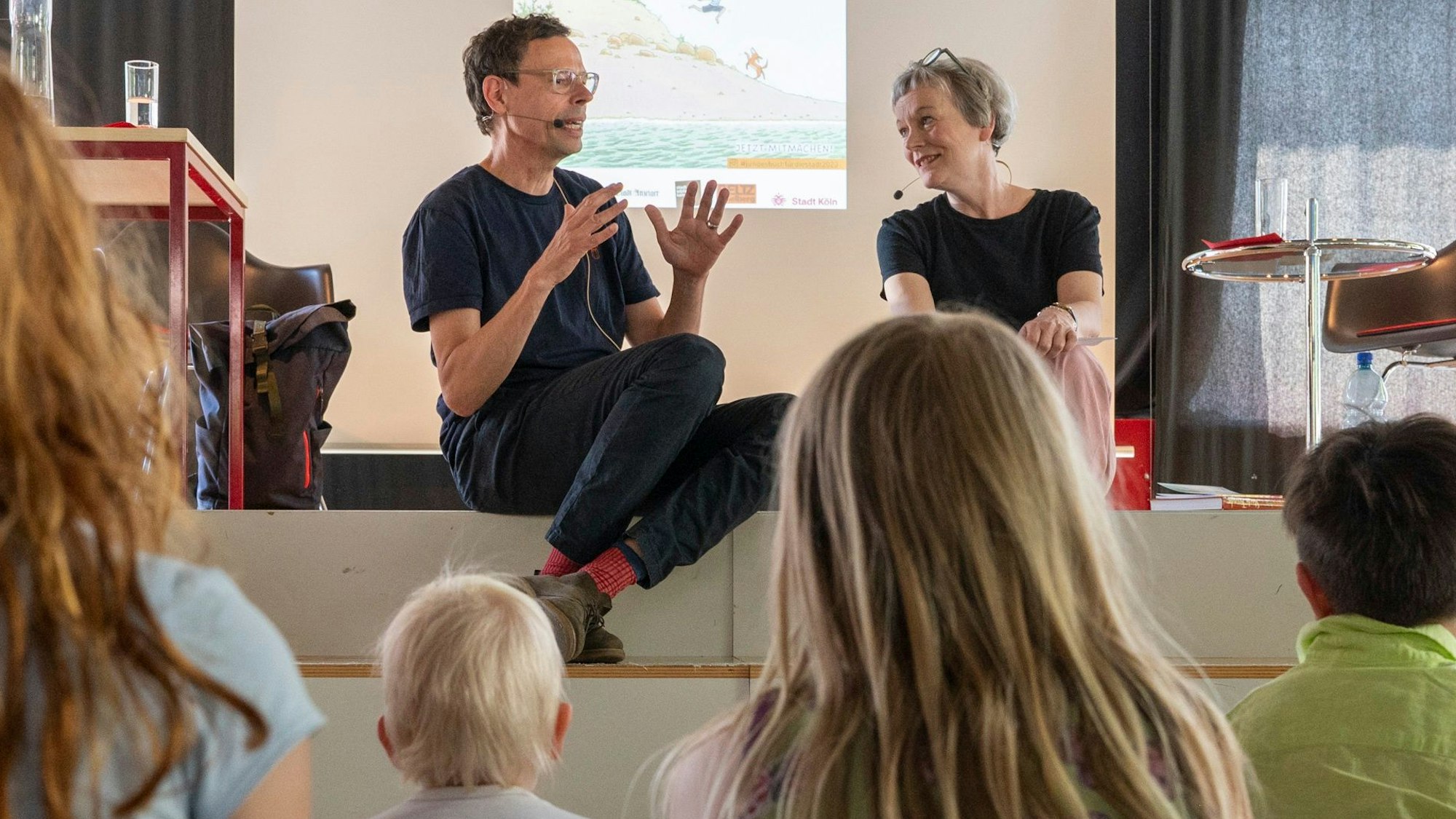 Philip Waechter sitzt mit Moderatorin Britta Weyers auf einer Bühne, davor sitzen Kinder.