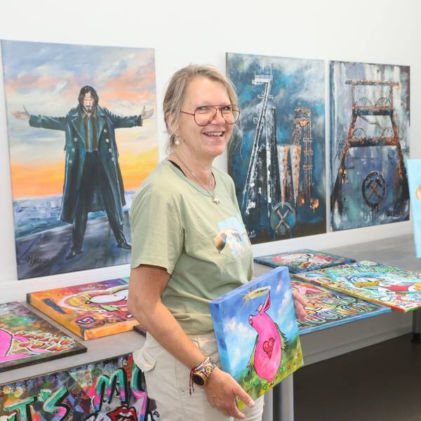 Eine Frau steht vor einer Bilderausstellung. Sie selbst hält ein Bild in der Hand, auf dem ein pinkes Schwein mit Herzkette um den Hals zu sehen ist.