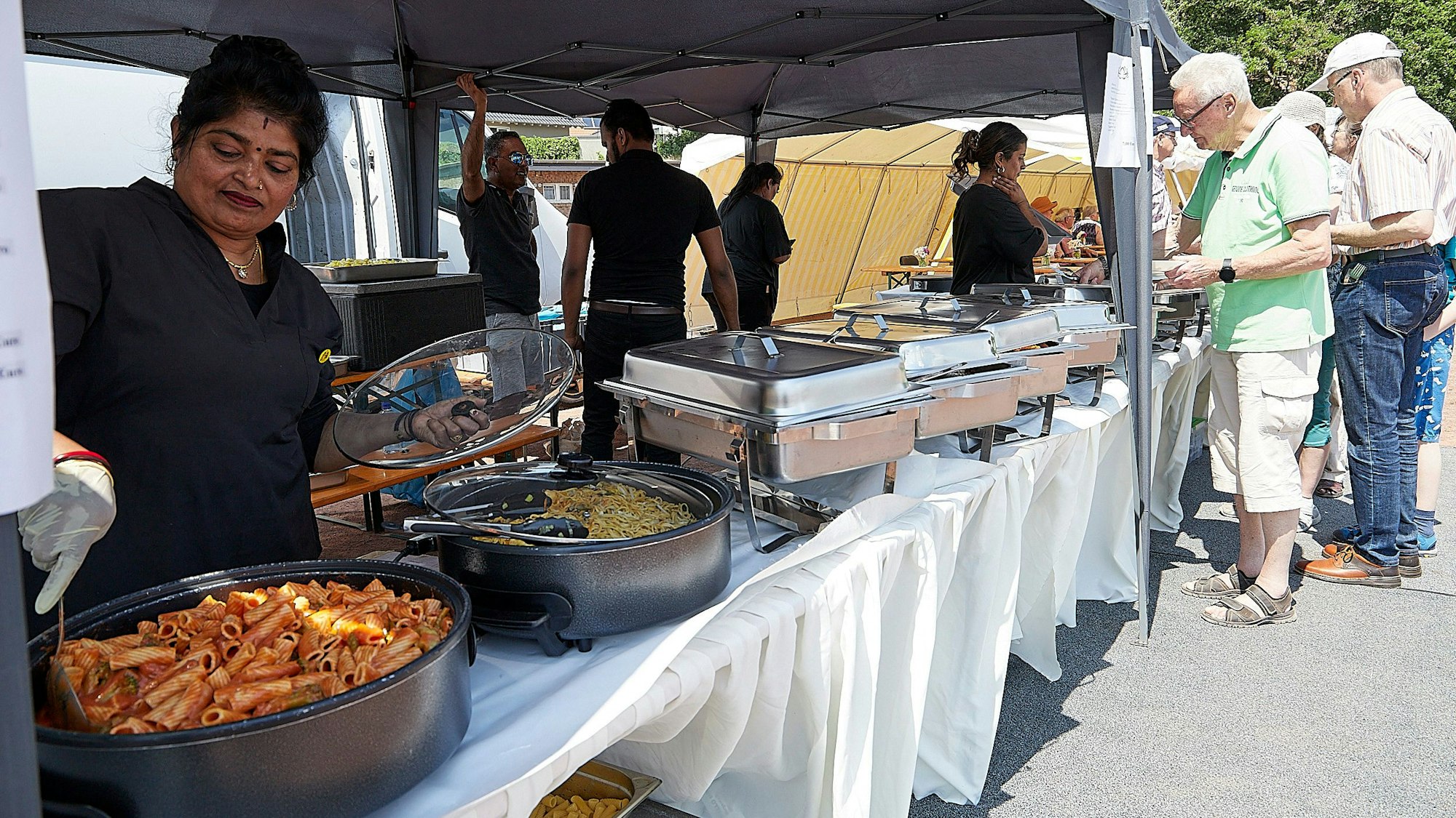 Das Bild zeigt eine Frau, die in Töpfen mit indischen Gerichten rührt. Im Hintergrund stehen Menschen am Buffet an.