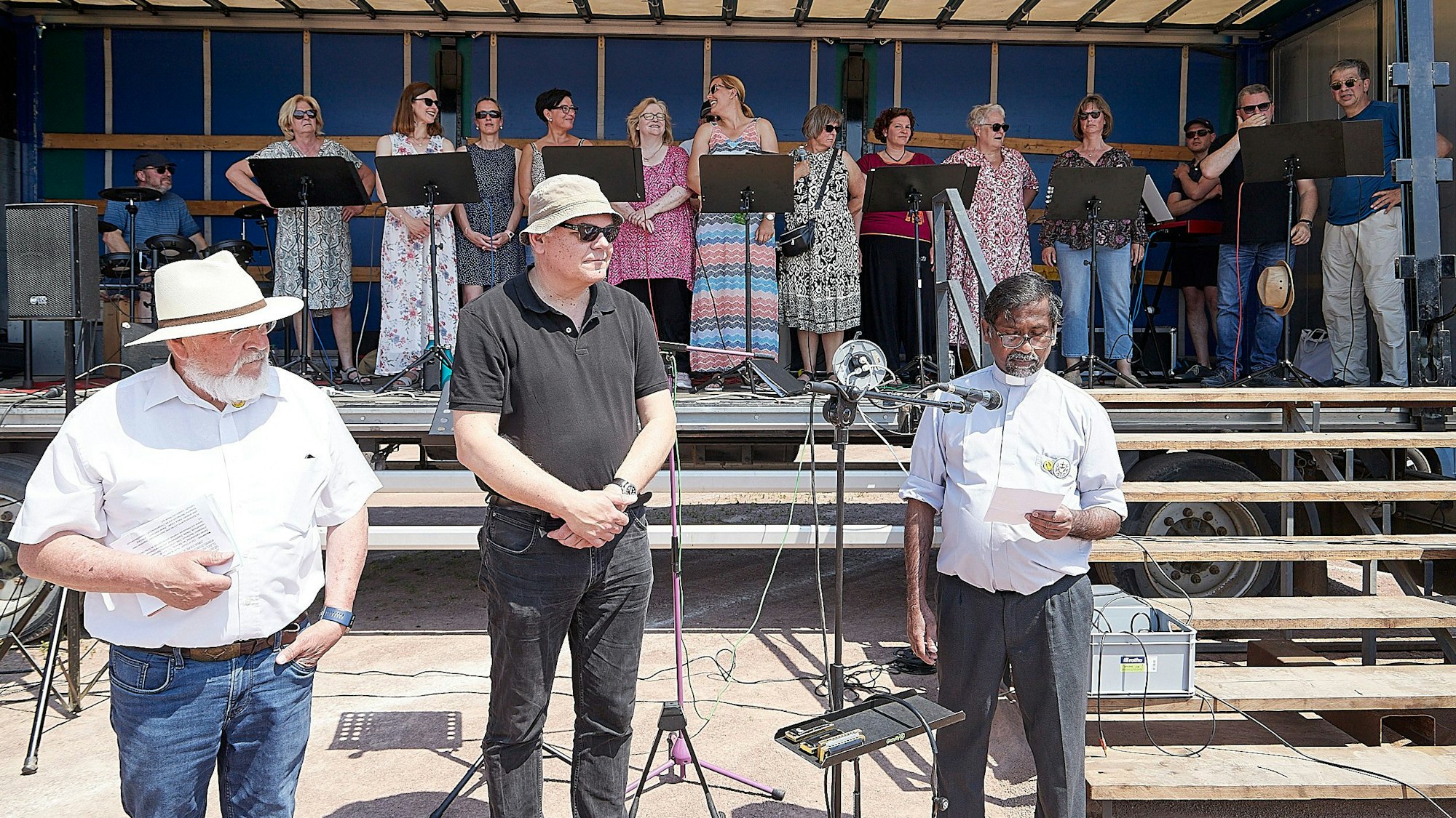 Das Bild zeigt drei Männer vor einer Bühne, auf der ein Chor steht.