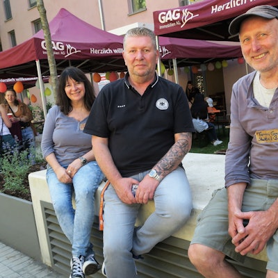 Eine Frau und zwei Männer sitzen vor einem Stand der GAG.